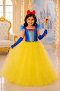 A young girl modeling a Snow White costume in a grand, castle-themed room. The costume features a bright yellow tulle skirt, a blue bodice with gold glitter details, blue gloves, a red bow headband, and a gold scepter.