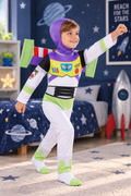 A young boy wearing a Buzz Lightyear space ranger costume in a child's bedroom. The costume includes a purple hood, white pants and sleeves, green accents, and a black chest panel with colorful details.