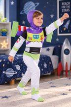 A young boy wearing a Buzz Lightyear space ranger costume in a child's bedroom. The costume includes a purple hood, white pants and sleeves, green accents, and a black chest panel with colorful details.
