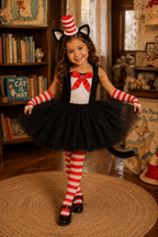 A young girl modeling a Cat in the Hat costume in a cozy, book-filled room. The costume includes a black tulle tutu, a white top with a red bow, red and white striped arm warmers and tights, a red and white striped hat with cat ears, and a black tail.