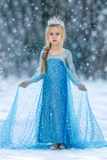 A young girl dressed as Elsa stands in a snowy outdoor setting wearing a sparkling blue gown with a matching tiara.