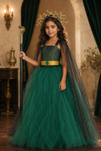 A young girl wearing a green and black tulle princess dress with a gold belt, a black sparkly cape, and a gold antler crown, holding a staff in a royal-themed room.