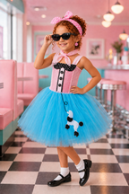 A young girl models a pink and blue poodle-themed costume in a retro-style pink diner. The costume features a pink corset top with black polka-dot trim and buttons, a bright blue tulle skirt with a black-and-white poodle applique, a pink bow headband, and black-and-white socks and shoes.