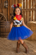 A young girl modeling a Woody-inspired cowboy costume in a barn setting. The costume features a cow-print bodysuit with a red bandana, a gold belt, a bright blue tutu skirt, and brown cowboy boots.