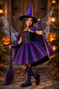 A young girl wearing a purple and black witch costume stands on outdoor stone steps decorated with cobwebs, jack-o'-lanterns, and lanterns. She holds a purple broom and wears a pointed purple hat, black-and-purple striped socks, and black shoes.
