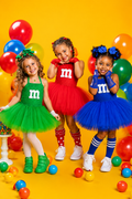Three young girls modeling colorful M&M-themed tutu dresses in green, red, and blue, standing on a bright yellow background with balloons and candy balls.