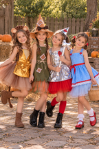 Four young girls wearing Wizard of Oz themed costumes pose together outdoors on a cobblestone path with pumpkins and a wooden fence in the background. The costumes include a lion, green witch, silver wizard, and blue-and-white dress with red accents.